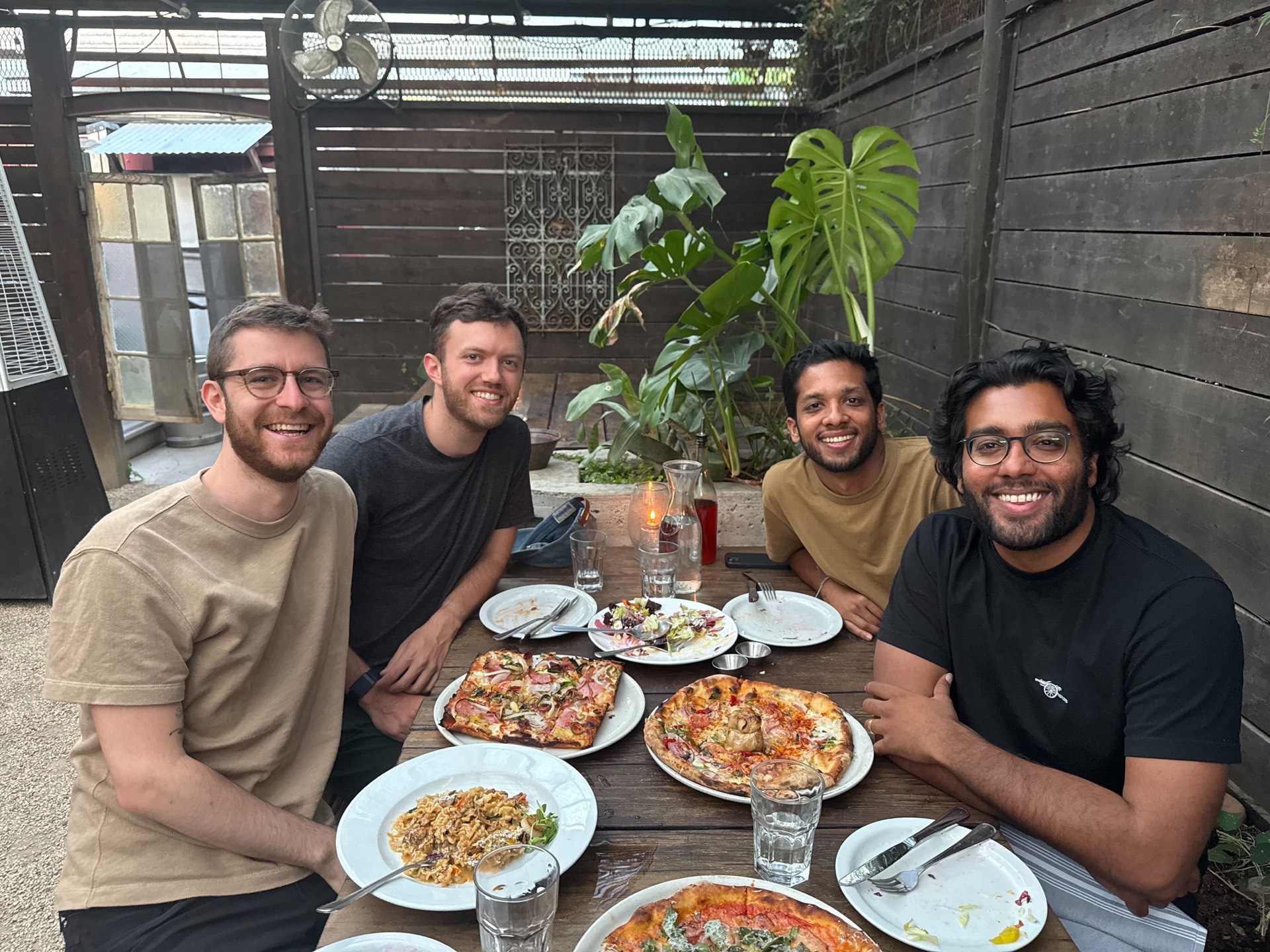 Four people laughing at an outdoor patio table — pizzas, pasta, drinks, wood fence, monstera.