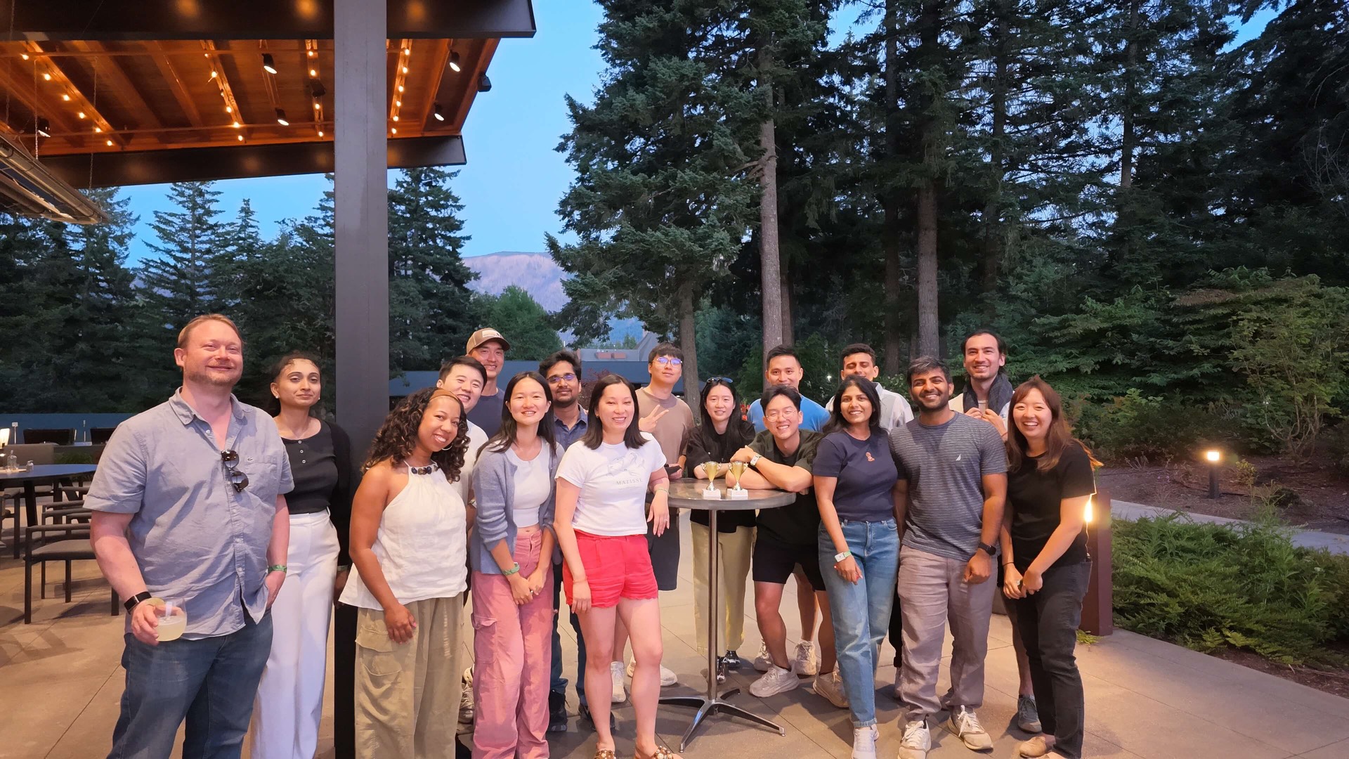 Raspberry AI team at dusk under a lit pavilion, string lights, forest behind.